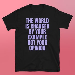 以下が含まれることがあります： 紫色の文字で「THE WORLD IS CHANGED BY YOUR EXAMPLE NOT YOUR OPINION」とプリントされた黒いTシャツ