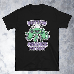 May include: Black t-shirt featuring a green controller design with tentacles and the words "BUTTON MASHER MUTANT" in white and purple. The controller has a smiling face and is a fun, playful design.