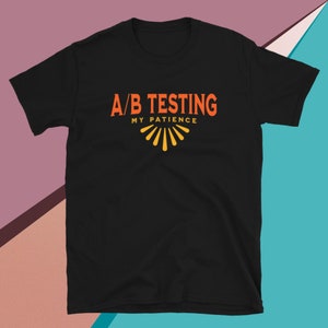 May include: Black t-shirt with the text "A/B Testing My Patience" in orange and yellow. The text is in a retro font style.