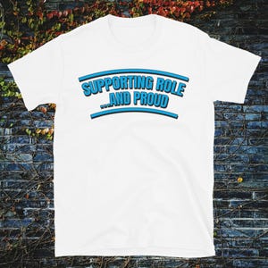 May include: White t-shirt with the text "SUPPORTING ROLE ...AND PROUD" in blue and black lettering. The text is arched across the chest. The shirt is short-sleeved.