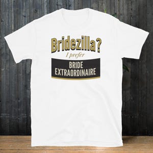 Puede incluir: Camiseta blanca con el texto "Bridezilla? I prefer Bride Extraordinaire" en dorado y negro. La camiseta tiene cuello redondo y mangas cortas. El texto está centrado en la parte delantera de la camiseta.