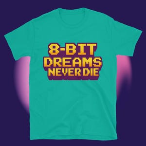 Puede incluir: Una camiseta turquesa con el texto "8-BIT DREAMS NEVER DIE" en letras pixeladas amarillas y moradas. La camiseta es de cuello redondo y manga corta.