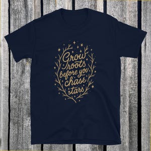 Puede incluir: Camiseta azul marino con el texto dorado "Grow roots before you chase stars" en un diseño floral. El diseño está centrado en la parte delantera. La camiseta está hecha de un material suave.