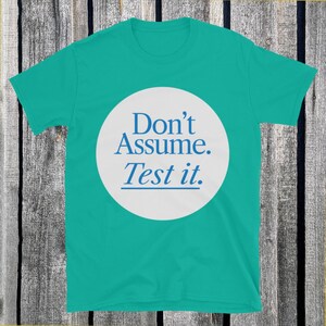 May include: A teal t-shirt with a white circle in the center. The circle contains the text "Don't Assume. Test it." in blue, sans-serif font. The shirt is made of a soft material.
