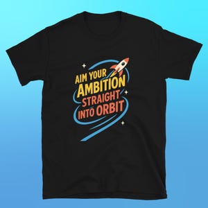 Könnte beinhalten: Schwarzes T-Shirt mit dem Text "AIM YOUR AMBITION STRAIGHT INTO ORBIT" in Gelb, Orange und Blau. Eine Cartoon-Rakete ist über dem Text abgebildet. Das T-Shirt hat einen Rundhalsausschnitt.