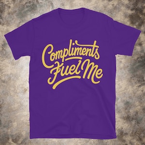 May include: A purple t-shirt with the text "Compliments Fuel Me" in a yellow, cursive font. The shirt is a solid color and has short sleeves.