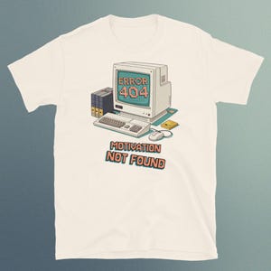 Può includere: T-shirt color crema con grafica di computer retrò. Il design presenta un monitor di computer vintage con la scritta "ERROR 404" e il testo "MOTIVATION NOT FOUND". La grafica include tastiera, mouse e floppy disk nei toni del verde acqua, arancione e beige.
