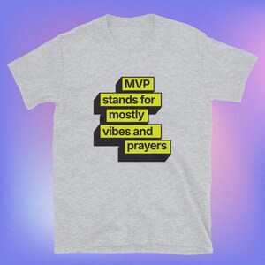 Op de afbeelding: Lichtgrijs t-shirt met de tekst "MVP stands for mostly vibes and prayers" in een zwart en geel ontwerp. Het shirt heeft korte mouwen en een ronde hals.