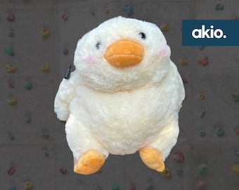 Pode incluir: Um pato de pelúcia branco com pés laranja e uma alça preta. O pato está sentado em uma parede texturizada cinza com agarras de escalada coloridas.
