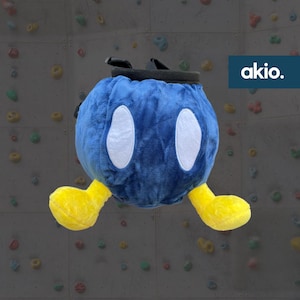 Puede incluir: Bolsa de magnesio de escalada de felpa azul con brazos amarillos y ojos blancos. La bolsa tiene una correa negra y la palabra "akio." está impresa en una etiqueta blanca.