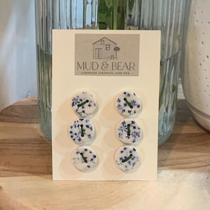 Peut inclure: Six boutons en céramique ronds avec une base blanche et un motif floral bleu, disposés en deux colonnes de trois sur une carte de présentation rectangulaire. La carte porte le texte MUD & BEAR HANDMADE CERAMICS, YASS NSW et une illustration.
