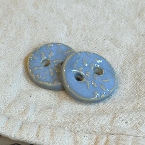 Puede incluir: Dos botones de cerámica de color azul claro con forma redonda, acabado brillante y un patrón texturizado en relieve en la superficie. Cada botón cuenta con dos orificios centrales para su fijación, mostrando un aspecto esmaltado y liso.