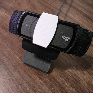 May include: Black Logitech HD 1080p webcam with a white privacy cover. The webcam has a black stand and a black and purple mesh design on the sides. The Logitech logo is visible on the front.
