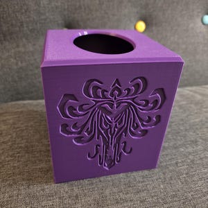 May include: A square, purple tissue box with a decorative, carved design on the front. The top has a circular opening for tissue dispensing. The box is made of a solid material and sits on a gray surface.