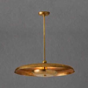 Puede incluir: Una lámpara colgante de color dorado, de estilo moderno de mediados de siglo. La lámpara tiene una pantalla circular y una fuente de luz más pequeña y redondeada debajo. Cuelga de una fina varilla fijada a un soporte de techo circular.