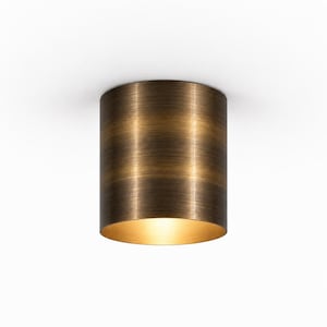May include: A cylindrical ceiling light fixture with a brushed bronze finish. The exterior features horizontal metallic bands with a textured appearance, while the interior is illuminated, casting a warm glow from the open bottom circular base.