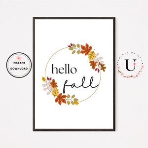 May include: A printable wall art design featuring a gold circle wreath with autumn leaves in shades of orange, red, yellow, and green. The text "hello fall" is written in black script font inside the wreath.