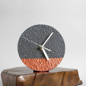 May include: A modern clock with a gray and copper face. The clock is sitting on a wooden base.