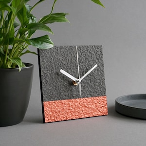 May include: A square clock with a black textured face and a copper colored bottom. The clock has white hands and a silver center.