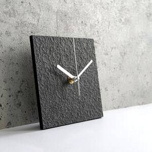 May include: A square gray clock with white hands and a gold center. The clock face has a textured surface.