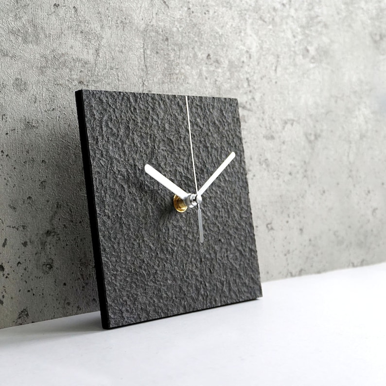 May include: A square gray clock with white hands and a gold center. The clock face has a textured surface.