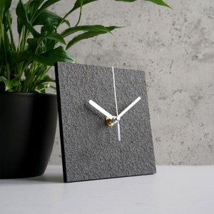May include: A gray square clock with white hands and a silver knob. The clock has a textured surface that resembles stone.