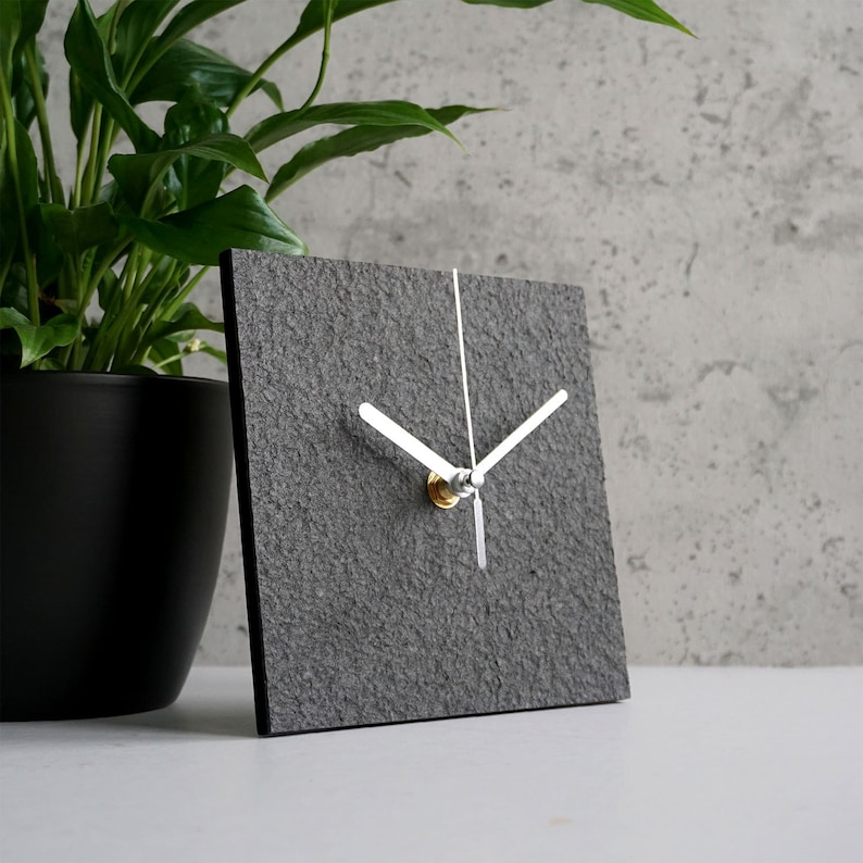 May include: A gray square clock with white hands and a silver knob. The clock has a textured surface that resembles stone.