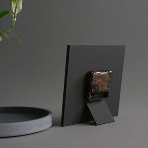 May include: Black square clock with a black stand. The clock face is not visible in the image. The clock is on a gray surface.