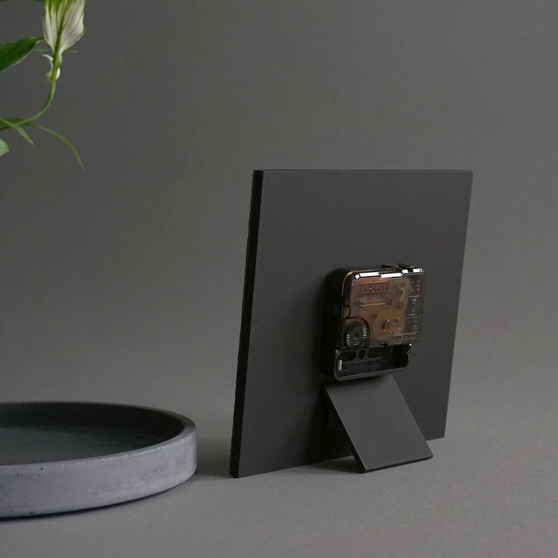 May include: Black square clock with a black stand. The clock face is not visible in the image. The clock is on a gray surface.