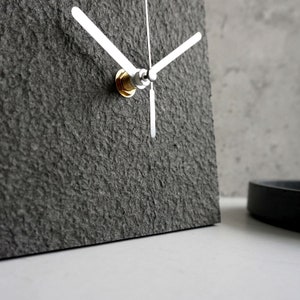 May include: A gray square clock with silver hands and a gold knob. The clock has a textured surface.