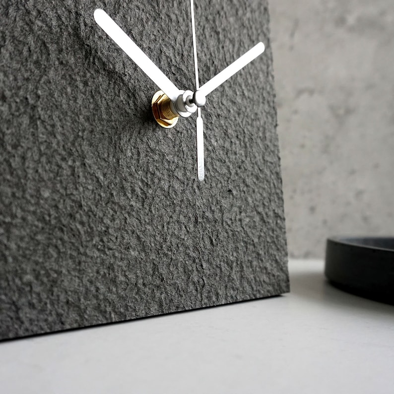 May include: A gray square clock with silver hands and a gold knob. The clock has a textured surface.