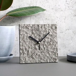 May include: Square clock with a textured, grey stone-like face and black hands. The clock is set on a grey surface, with a potted plant and a small tray in the background. The clock's design is minimalist and modern.