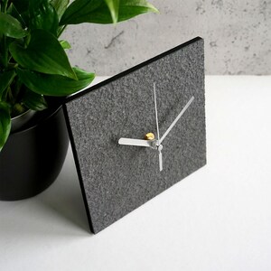 May include: A gray felt clock with silver hands and a gold center. The clock is square and has a simple design.