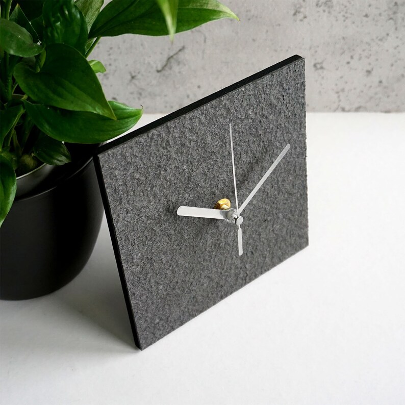 May include: A gray felt clock with silver hands and a gold center. The clock is square and has a simple design.