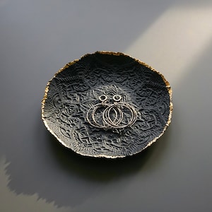 Può includere: Piatto portaoggetti in ceramica nero e oro con un design a mandala. Il piatto ha un bordo dorato ed è decorato con un motivo in rilievo. Due orecchini a cerchio dorati sono appoggiati sul piatto.