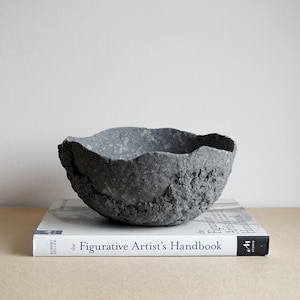 Könnte beinhalten: Eine handgefertigte, graue Schale mit strukturierter Oberfläche und unebenem Rand. Die Schale steht auf einem Buch mit dem Titel "The Figurative Artist's Handbook". Die raue Textur der Schale kontrastiert mit dem glatten Buchumschlag.