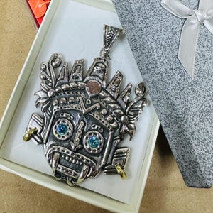 Puede incluir: Un colgante plateado con un diseño de rostro estilizado y detallado, presentado en una caja blanca. El colgante tiene ojos de piedra azul y detalles dorados. Se ve un paño rojo con el texto "MARASHINE" y "Paño Maravilloso", junto con un recuerdo de "TAXCO".