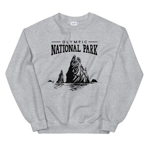May include: A light gray sweatshirt featuring a black graphic of a mountain range and the text "OLYMPIC NATIONAL PARK". The design includes a depiction of the park's natural landscape, with a mountain and water elements.