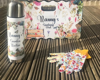 May include: A gardening gift set with a white and floral patterned thermos, a white and floral patterned kneeling pad with the text "Nanny's Gardening Pad", and a pair of white and floral patterned gardening gloves.