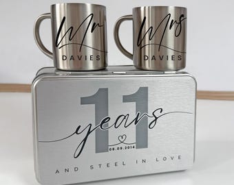 May include: Two stainless steel mugs with handles, one labeled "Mr" and the other "Mrs", both with the last name "Davies" in black script. They sit atop a silver box with "11 years" and "And Steel in Love" printed on it.