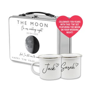 Op de afbeelding: Een zilveren metalen lunchbox met een wit label waarop staat "THE MOON On our wedding night 10/04/2015 And I'm still over the moon HAPPY 'TIN' YEAR" met een zwart-witte afbeelding van de maan. Twee witte emaille mokken met zwarte tekst die "Jack" en "Sarah" zegt met een hartje naast elke naam.