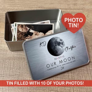 Op de afbeelding: Een zilveren metalen fotoblik met een deksel met een maangrafiek en de woorden "OUR MOON". De blik bevat verschillende zwart-wit foto's. Tekst op het deksel bevat namen en een datum. Een rood hart zegt "PHOTO TIN!"
