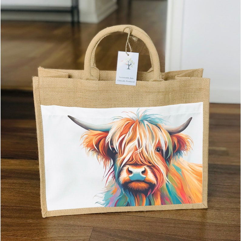 May include: A brown jute tote bag with a white canvas insert featuring a colorful illustration of a Highland cow. The bag has two handles and a tag that reads "Sustainable Joy Ethically Produced".