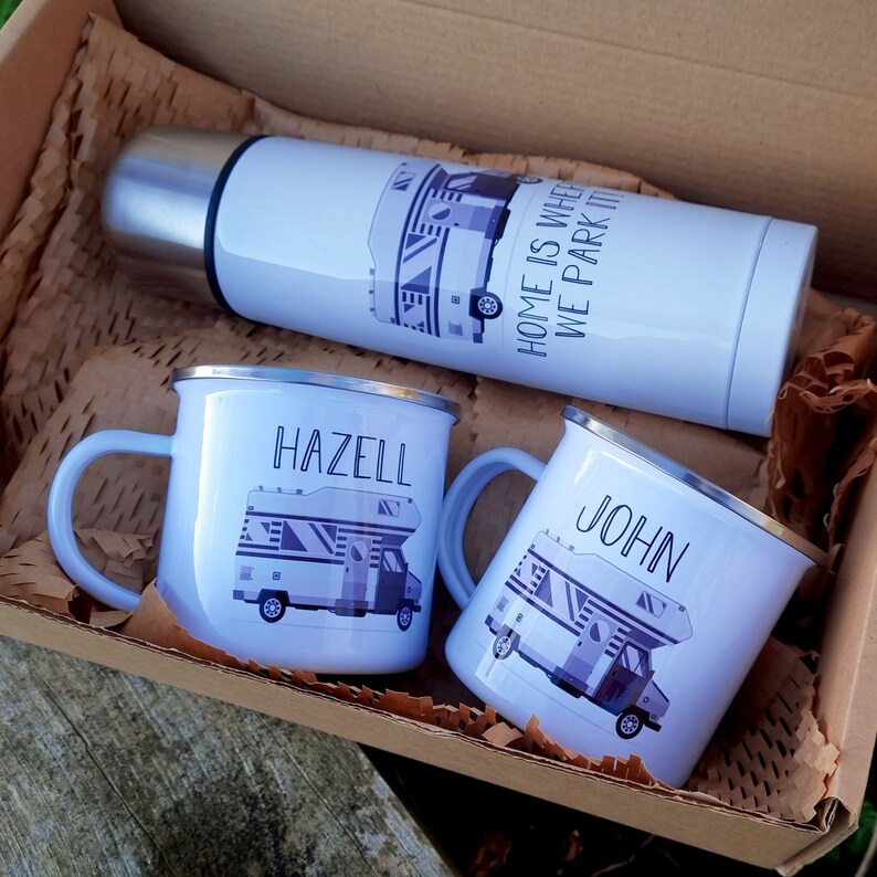 May include: Two white enamel mugs with personalized names "Hazell" and "John" and a camper van graphic. A white thermos with a camper van graphic and the text "Home is where we park it" is also included. The mugs and thermos are in a brown cardboard box with brown packing paper.