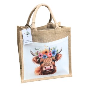 May include: A brown jute tote bag with a white pocket featuring a watercolor illustration of a Highland cow with a floral crown. The text "Sustainable Jute Officially, Proudly" is printed on a tag attached to the bag.