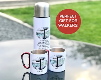 May include: Two white ceramic mugs with a red carabiner handle and a stainless steel thermos. The mugs and thermos have a black and white illustration of a signpost with the text "NO NEED TO RUN SLOW DOWN THE FACE BREATHE IN FRESH AIR FEEL THE SUN ON YOUR FACE AND WHEN YOU SEE A NICE SPOT FOR A BREW WHILE WALKING" and the names "NIKKI" and "JOOLS" printed on the mugs. The text "PERFECT GIFT FOR WALKERS!" is printed on a red circle.