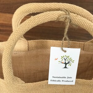 May include: A brown jute tote bag with white fabric lining. The bag has two thick rope handles and a small white tag that reads "Sustainable Jute Ethically Produced" with a small tree and bird illustration.