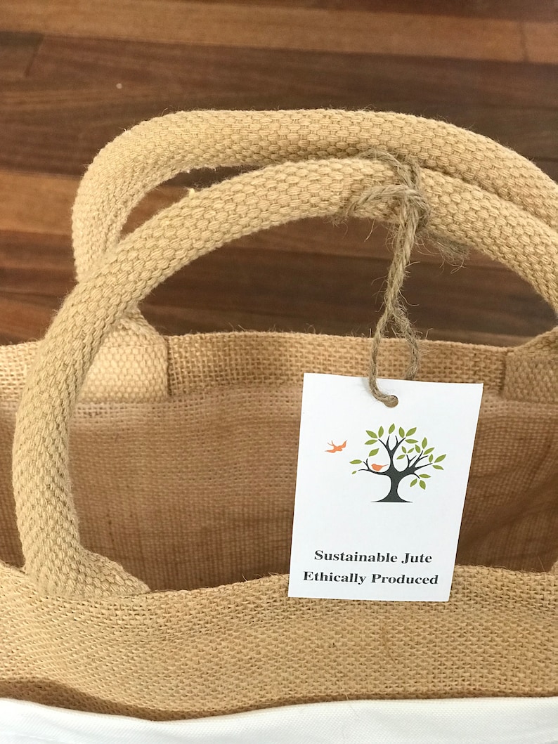 May include: A brown jute tote bag with white fabric lining. The bag has two thick rope handles and a small white tag that reads "Sustainable Jute Ethically Produced" with a small tree and bird illustration.