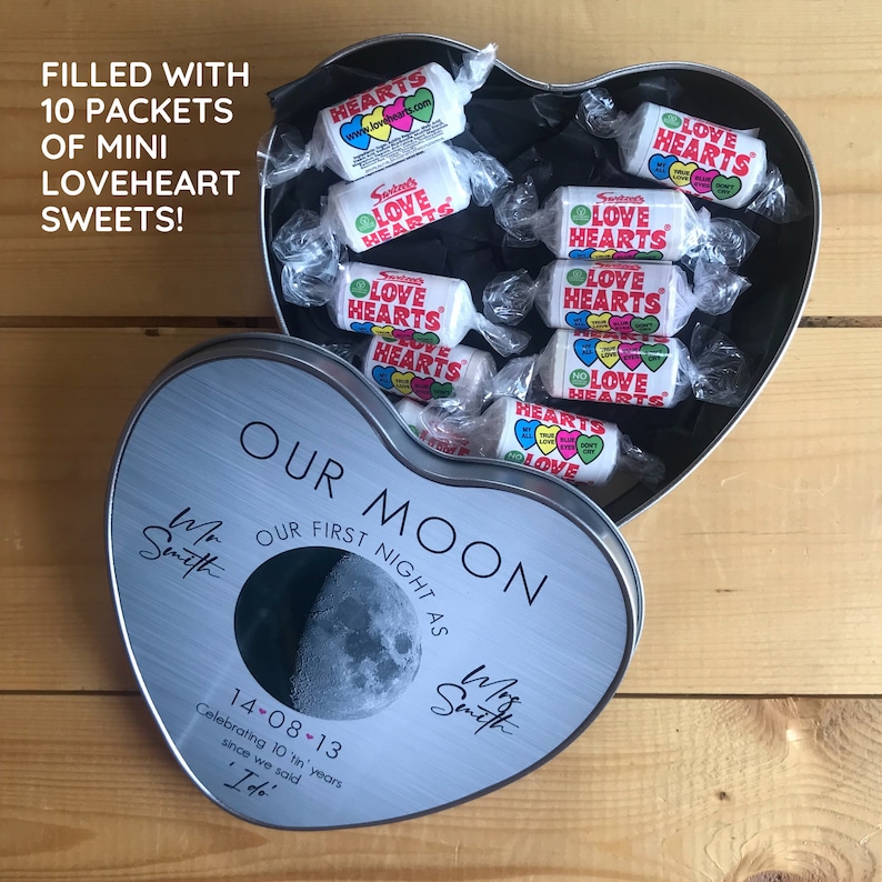 May include: A silver heart-shaped tin filled with 10 packets of Love Heart candies. The tin has a moon design and the text "Our Moon Our First Night As Mr. & Mrs. Smith 14-08-13 Celebrating 10 'tin' years since we said I do."