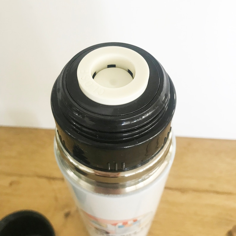 May include: Close-up of the top of a stainless steel thermos with a black lid and a white plastic stopper. The stopper has a small hole in the center.